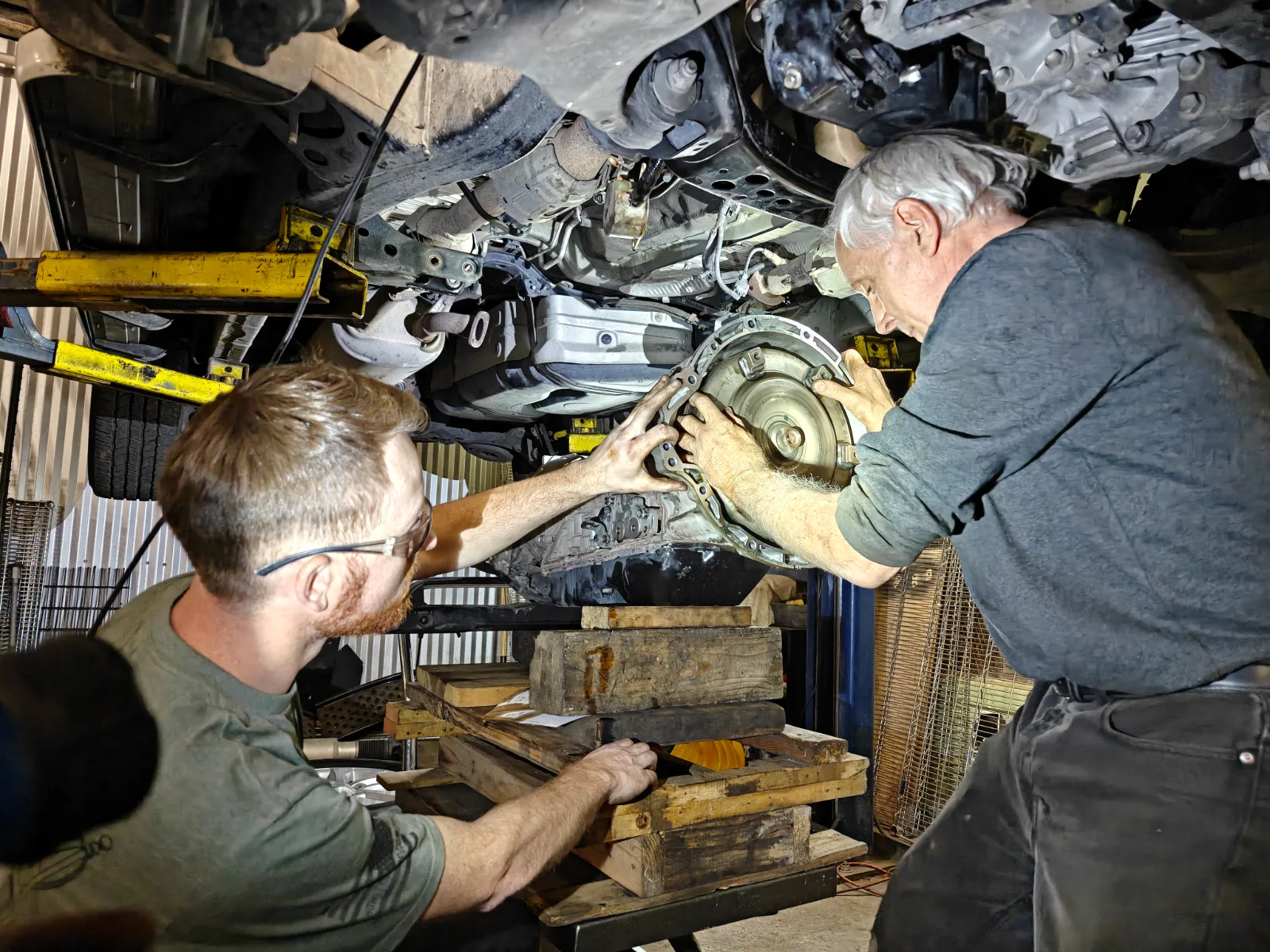 Ed and Matt underneath a car on the lift, together guiding a torque converter into the bellhousing