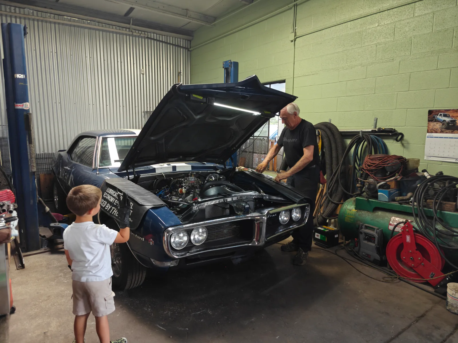 Ed working under the raised hood of a dark blue 1960s Pontiac Firebird in the shop while a young boy watches