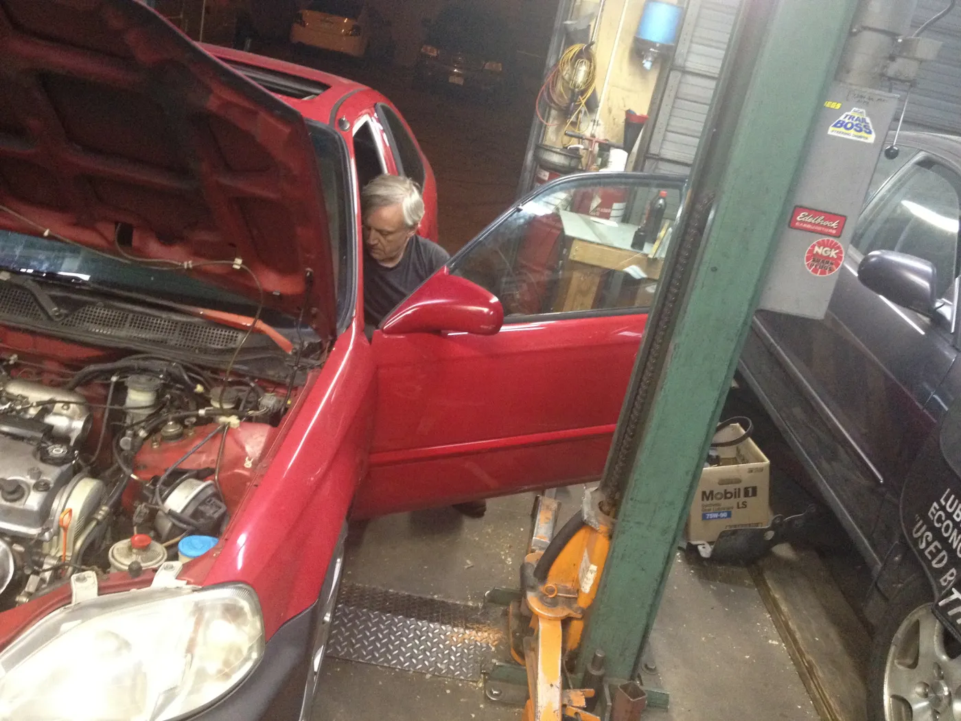 Ed reaching into the engine bay of a red coupe raised on the lift inside the shop